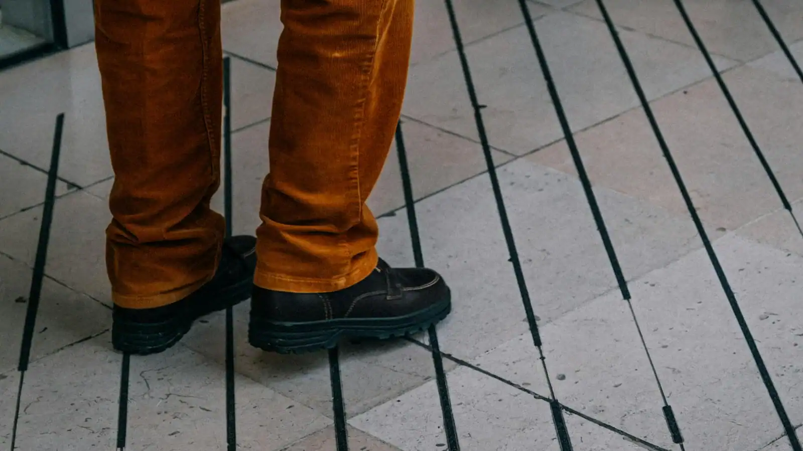 Close-up of feet wearing brown pants and black shoes on a tiled floor.