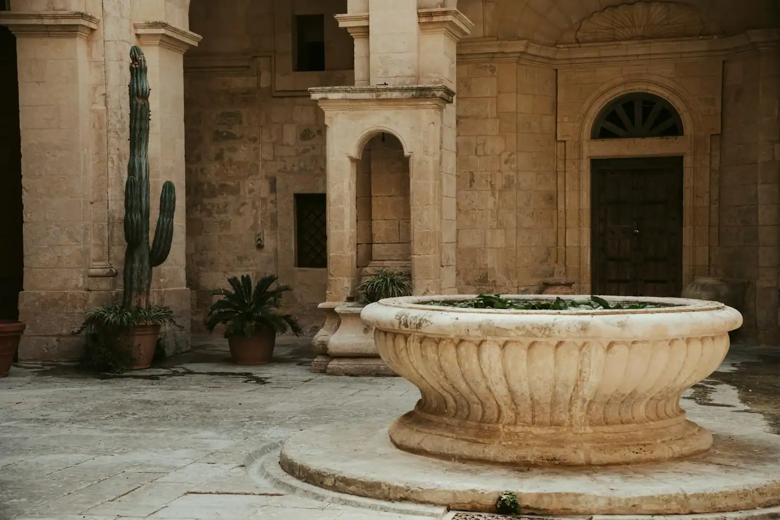Stone courtyard in Malta featuring a large central fountain and potted plants.