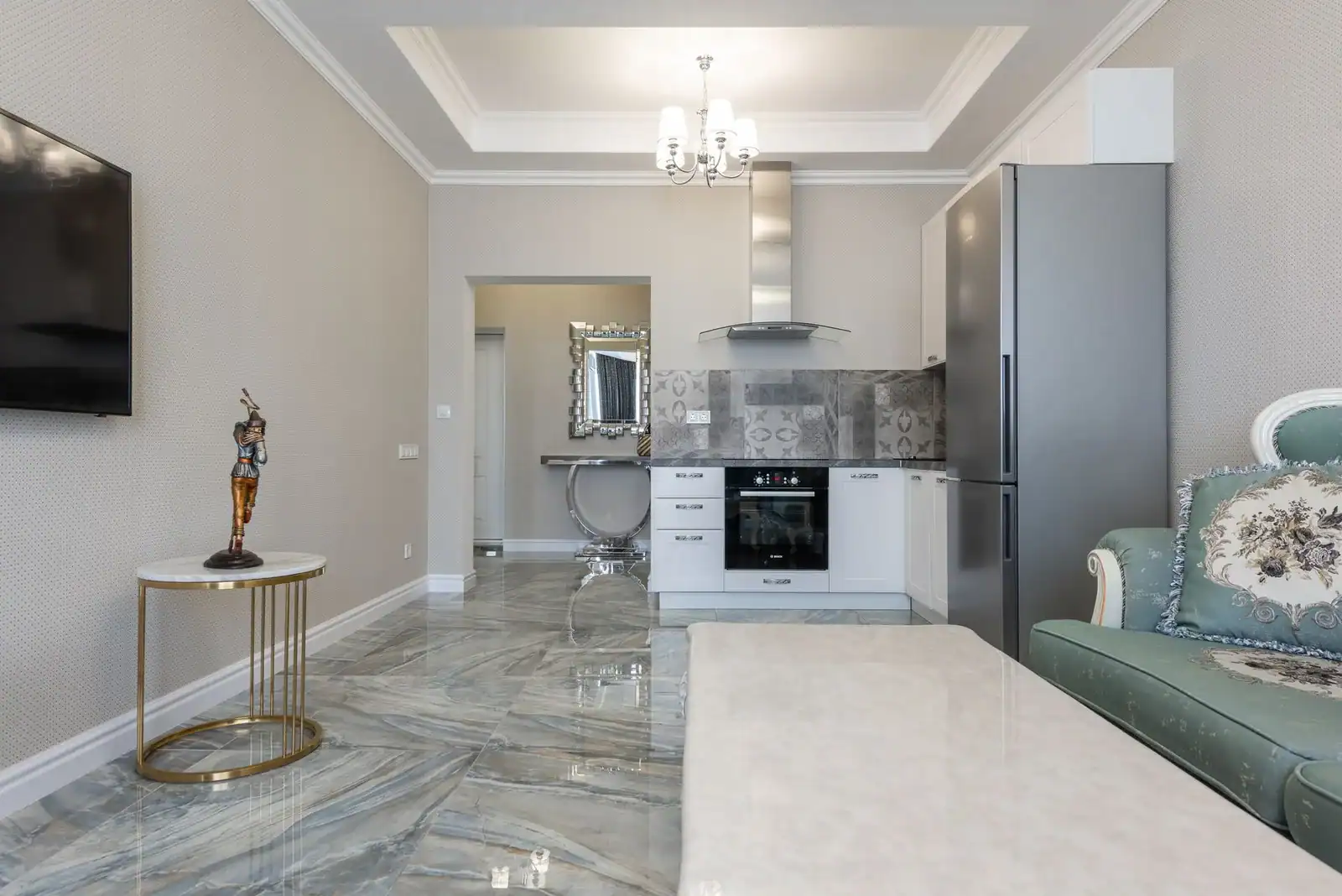 Contemporary kitchen and living room interior with sofa against tables and electric appliances under ceiling with chandelier in house
